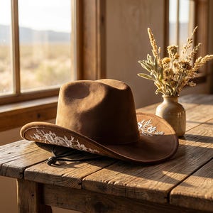 Op de afbeelding: Een bruine vilten cowboyhoed met witte bloemenborduursels langs de rand en een zwarte kinband met een verstelbare sluiting. De hoed heeft een gestructureerde kroon en een brede, gebogen rand met gedetailleerd stikwerk aan de buitenkant.