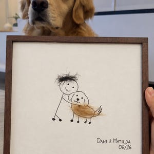 May include: A square framed artwork featuring a black ink stick figure drawing of a person hugging a dog. The person has black hair made of real hair, and the dog has a golden-brown fur body. The text "DANI & MATILDA 06/26" is written in black ink.