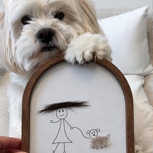 May include: An arched wooden frame containing a white background with a black ink stick figure drawing of a person holding a leash attached to a dog. The person has a dark hair accent, and the dog has a textured beige fur accent. Text reads Dani & Matilda 06/26.