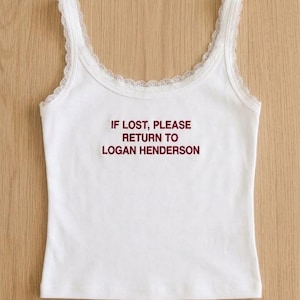 Puede incluir: Una camiseta de tirantes blanca sin mangas con escote redondo y un delicado borde de encaje a lo largo de los tirantes y el cuello. El centro del pecho presenta un texto bordado en rojo oscuro que dice: IF LOST, PLEASE RETURN TO LOGAN HENDERSON.