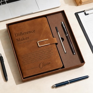 May include: A brown notebook with a gold-toned metal clasp and the text Difference Maker, a definition, and the name Olivia engraved on the cover. The notebook is inside a brown gift box alongside a matching pen and a thin metal stylus.