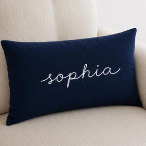 Peut inclure: Un coussin rectangulaire bleu marine avec le prénom "sophia" brodé en écriture cursive blanche au centre. Le coussin présente une surface en tissu uni et lisse, et il est positionné horizontalement sur un fond de couleur neutre.