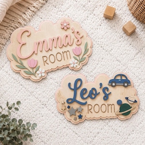 Op de afbeelding: Twee wolkvormige houten wandborden met verhoogde letters. Het ene bord toont de naam Emma's ROOM in roze met bloemaccenten. Het andere bord toont de naam Leo's ROOM in blauw met een auto, sterren en planeetmotieven bevestigd.