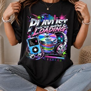 May include: A black t-shirt featuring a graphic print with the text "PLAYLIST LOADING" and "2000s VIBES". The design includes a blue digital music player, a stack of colorful compact discs, silver headphones, and a loading bar with music icons.