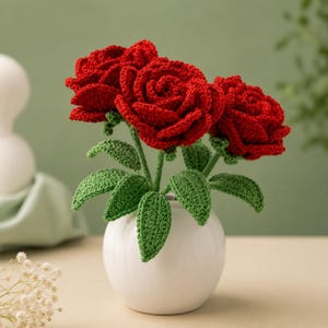 May include: Three crocheted red roses with textured green leaves and stems arranged in a small, smooth white ceramic vase. The flowers feature intricate yarn loops forming layered petals, while the foliage displays a detailed stitched pattern.