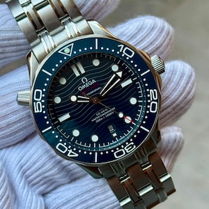 Op de afbeelding: Een roestvrijstalen Omega Seamaster Professional horloge met een blauwe keramische ring en een blauwe wijzerplaat met golfpatroon. Het horloge heeft lichtgevende uurmarkeringen, een datumvenster op de 6-uurpositie en een metalen schakelarmband.