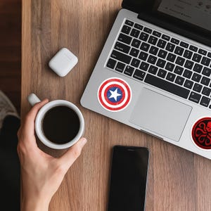 May include: A silver laptop featuring a circular sticker of a red, white, and blue shield with a central white star, and a second circular sticker with a red and black octopus-like emblem. A white mug of black coffee and a smartphone are nearby.