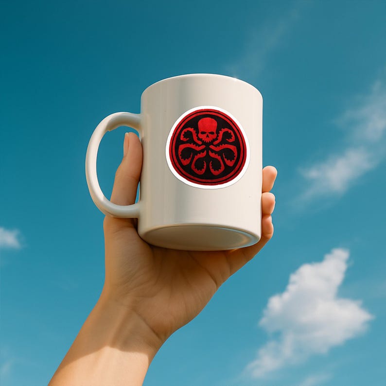 May include: A white ceramic mug featuring a circular graphic on the front. The graphic displays a red skull centered within a black and red octopus-like tentacle design, all enclosed within a thick red circular border against the white mug surface.