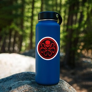May include: A matte blue water bottle featuring a circular sticker on the front. The sticker displays a red skull with tentacles extending downward, all enclosed within a red and white circular border against a dark background.