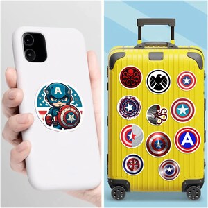 May include: A white smartphone case featuring a circular sticker of a cartoon superhero with a shield, and a bright yellow rolling suitcase decorated with various circular stickers including shield emblems, a skull logo, and a bird insignia.