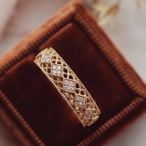 May include: A gold band ring featuring an intricate openwork lattice pattern across the entire surface. Small, clear, sparkling stones are set within the diamond-shaped cutouts of the metalwork, creating a textured and detailed appearance.