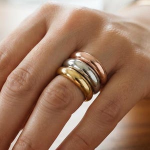 May include: A set of three stacked rings featuring a smooth, rounded profile. The rings are crafted in three distinct metallic finishes: a warm rose gold, a bright silver, and a rich yellow gold, all worn together on a single finger.