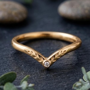 May include: A gold V-shaped ring featuring a delicate engraved leaf pattern along the band and a single small round white diamond set at the center point. The metal has a polished finish, and the design follows a chevron silhouette.