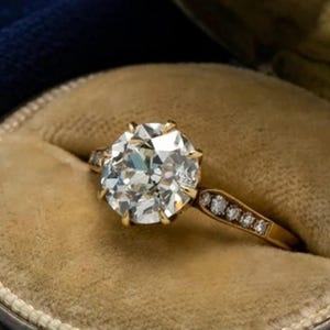 Puede incluir: Un anillo de oro con una gran piedra central redonda y facetada sujeta por seis garras, con una banda adornada por una hilera de pequeñas piedras claras de talla redonda a lo largo del aro, presentado en un joyero redondo forrado de terciopelo beige.
