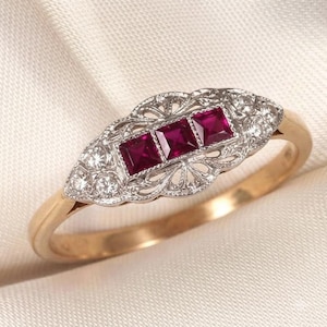 May include: A gold ring featuring a central horizontal row of three square-cut red rubies set in a white metal filigree mounting. The filigree design is accented with small round white stones on both sides of the rubies, set on a gold band.