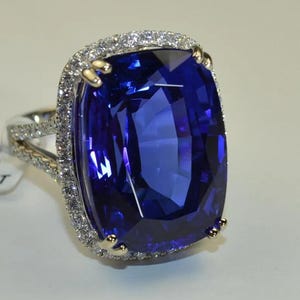 May include: A ring featuring a large, rectangular cushion-cut deep blue gemstone as the centerpiece. The stone is surrounded by a halo of small, clear, round-cut white stones. The band is split and encrusted with additional small white stones.