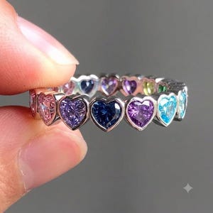 May include: A silver-toned eternity band featuring a continuous row of heart-shaped gemstones in various colors, including pink, purple, blue, and green. Each stone is set in a metallic frame, creating a vibrant, multi-colored circular ring design.