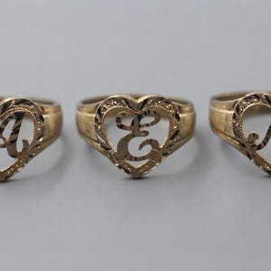 May include: Three gold-toned rings, each featuring a heart-shaped frame with a textured, diamond-cut border. Centered within each heart is a single, stylized capital letter: A, E, and M. The bands have a smooth, brushed finish on the sides.