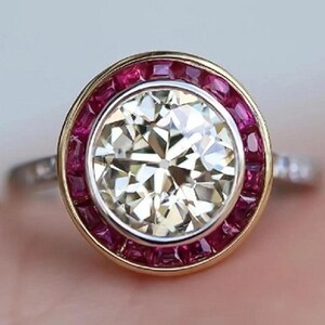 Könnte beinhalten: Ein Ring mit einem großen, runden, facettierten weißen Mittelstein in einer silberfarbenen Fassung. Der Mittelstein ist von einem Kranz aus kleinen, quadratisch geschliffenen, dunkelroten Steinen in einem goldfarbenen Rahmen umgeben.