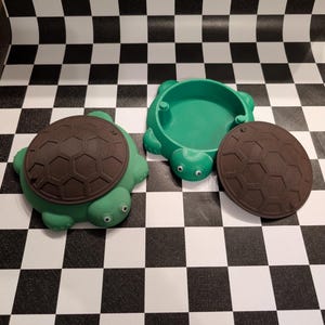 May include: Two turtle-shaped containers, one closed and one open with its brown shell lid removed. Each container has a bright green body with a textured brown shell top and small googly eyes attached to the head. The containers are hollow.