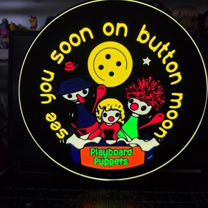 May include: A circular graphic display on a laptop screen featuring a yellow button icon, three stylised puppet characters, and the text "see you soon on button moon" in yellow, with "Playboard Puppets" in green text on an orange rectangular base.