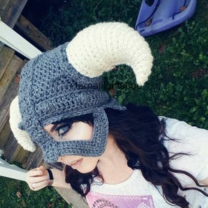 Puede incluir: Un gorro de crochet gris y blanco con forma de casco con cuernos. El gorro lo lleva puesto una persona con el pelo largo y oscuro. El gorro tiene una m&aacute;scara facial con un recorte para los ojos.