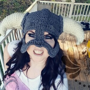 Puede incluir: Primer plano de una persona que lleva un casco de ganchillo gris con cuernos blancos. El casco tiene una m&aacute;scara facial con agujeros para los ojos y una expresi&oacute;n feroz. La persona sonr&iacute;e y mira directamente a la c&aacute;mara.