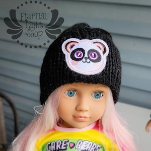 May include: A black knit beanie featuring a panda bear patch with pink and purple accents. The beanie is worn by a doll with blue eyes and pink and white hair. The doll is wearing a Care Bears shirt.