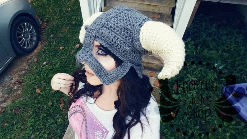 Puede incluir: Un gorro de crochet gris y blanco con cuernos. El gorro tiene forma de casco y un dise&ntilde;o detallado. El gorro lo lleva puesto una persona con el pelo largo y oscuro.