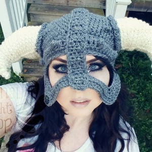 Puede incluir: Un casco gris de ganchillo con cuernos blancos. El casco tiene una m&aacute;scara facial con agujeros para los ojos. La persona que lleva el casco tiene el pelo oscuro y mira a la c&aacute;mara.