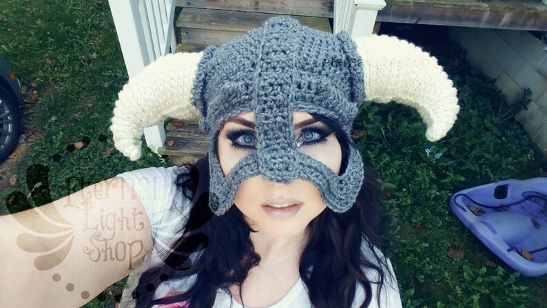 Puede incluir: Un casco gris de ganchillo con cuernos blancos. El casco tiene una m&aacute;scara facial con agujeros para los ojos. La persona que lleva el casco tiene el pelo oscuro y mira a la c&aacute;mara.