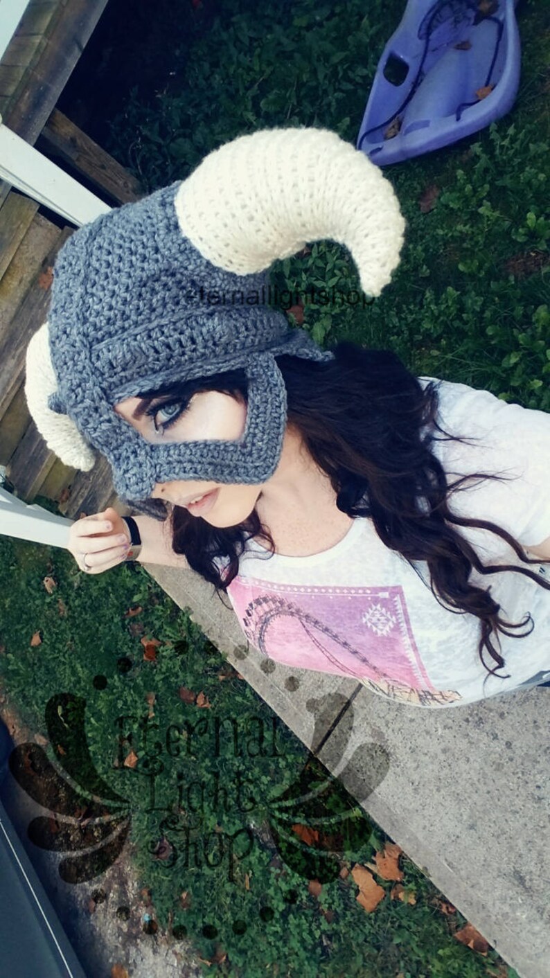 Puede incluir: Un gorro de crochet gris y blanco con forma de casco con cuernos. El gorro lo lleva puesto una persona con el pelo largo y oscuro. El gorro tiene una m&aacute;scara facial con un recorte para los ojos.