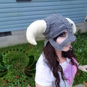 Puede incluir: Un gorro de crochet gris y blanco con cuernos, que se asemeja a un casco. El gorro tiene un dise&ntilde;o de m&aacute;scara facial con un agujero negro para el ojo. El gorro lo lleva una persona con el pelo largo y casta&ntilde;o.
