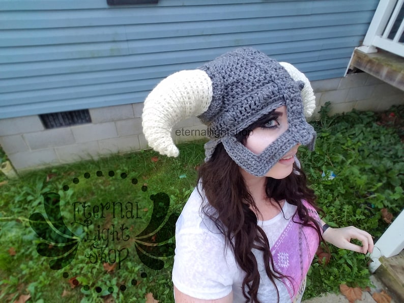 Puede incluir: Un gorro de crochet gris y blanco con cuernos, que se asemeja a un casco. El gorro tiene un dise&ntilde;o de m&aacute;scara facial con un agujero negro para el ojo. El gorro lo lleva una persona con el pelo largo y casta&ntilde;o.