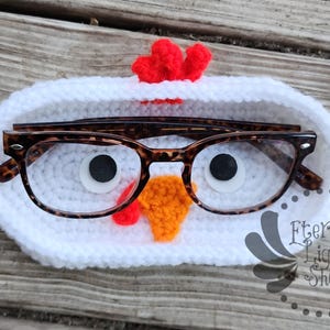 May include: A white crocheted eyeglass holder shaped like a chicken, with black and white eyes, an orange beak, and a red comb. Tortoiseshell eyeglasses rest inside the holder. The holder is on a wooden surface.