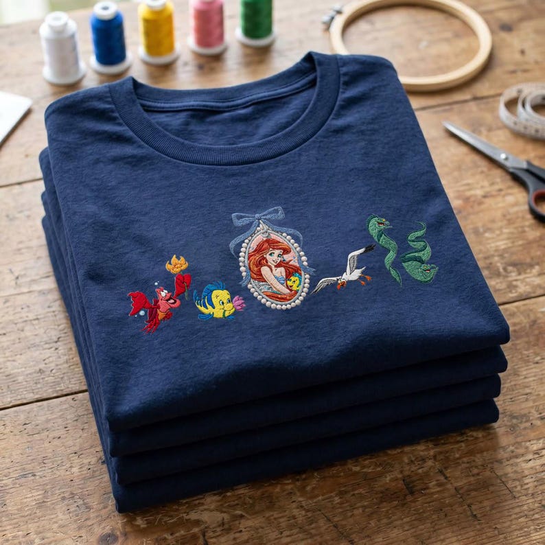 May include: A navy blue t-shirt featuring embroidered characters from The Little Mermaid, including a red crab, a yellow and blue fish, a central oval portrait of a mermaid with a blue bow, a white seagull, and two green eels.