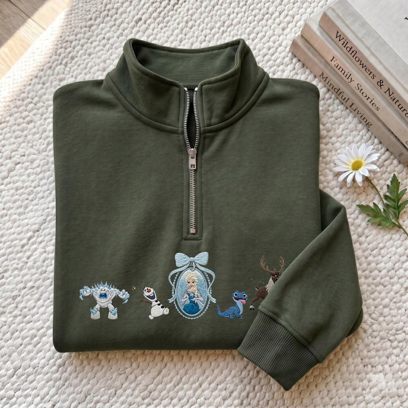 May include: An olive green quarter-zip sweatshirt featuring embroidered characters from the movie Frozen, including a snow monster, a snowman, a framed portrait of a queen, a small blue lizard, and a reindeer, all arranged across the chest.