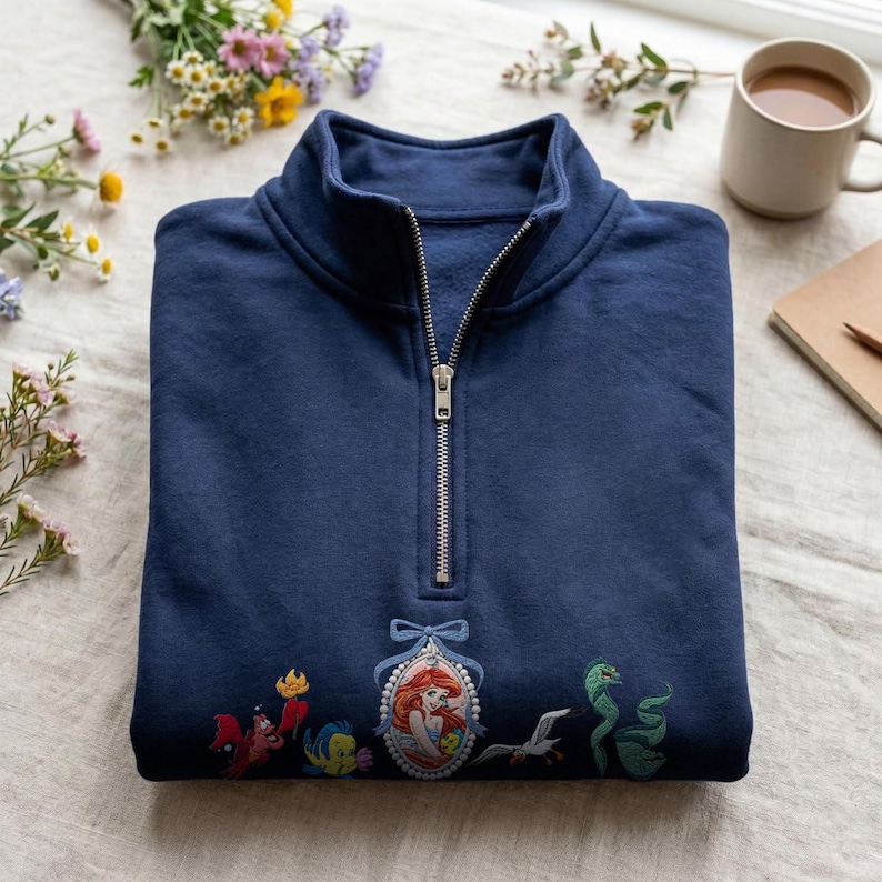 May include: A navy blue quarter-zip sweatshirt featuring an embroidered design of a central oval cameo portrait of a red-haired mermaid character, flanked by a red crab, a yellow fish, a white seagull, and a green eel, all with a blue bow.