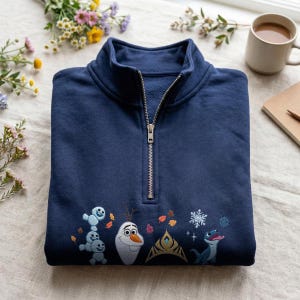 May include: A navy blue quarter-zip sweatshirt featuring embroidered characters and motifs including small snow creatures, a snowman, a golden crown, a blue lizard, and snowflakes, accented with scattered orange and brown autumn leaves.