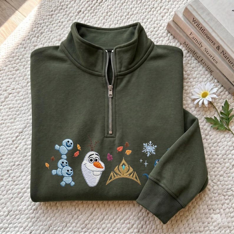 May include: An olive green quarter-zip sweatshirt featuring embroidered designs of a snowman, small snow creatures, a golden crown, and snowflakes. The embroidery includes colorful autumn leaves scattered around the characters and symbols on the front.