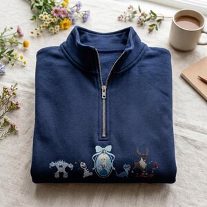May include: A navy blue quarter-zip sweatshirt featuring embroidered characters from the movie Frozen, including a snow monster, a snowman, a central portrait of a queen in a blue bow frame, a small blue lizard, and a reindeer with a red bird.