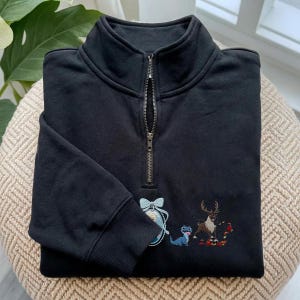 May include: A black quarter-zip sweatshirt featuring a high collar and a metal zipper. The front displays an embroidered oval portrait of a blonde character with a light blue bow, a small blue lizard, and a brown reindeer standing among red leaves.