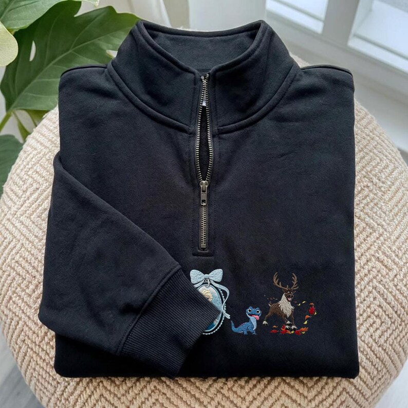 May include: A black quarter-zip sweatshirt featuring a high collar and a metal zipper. The front displays an embroidered oval portrait of a blonde character with a light blue bow, a small blue lizard, and a brown reindeer standing among red leaves.