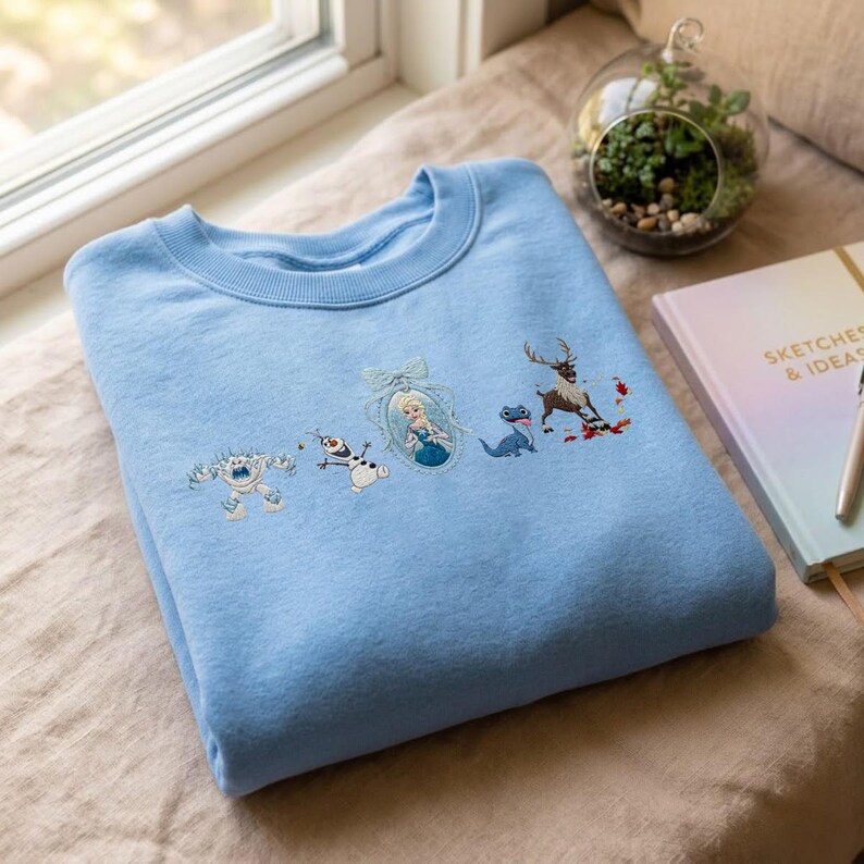 May include: A light blue crewneck sweatshirt featuring embroidered characters from the movie Frozen. The design includes a white yeti, a snowman, a female figure in a blue dress inside an oval frame, a small blue lizard, and a brown reindeer.