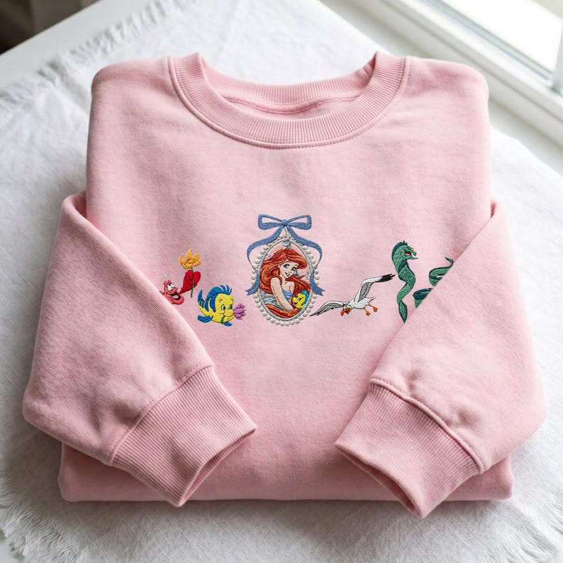May include: A light pink crewneck sweatshirt featuring embroidered characters from The Little Mermaid. The design includes a central portrait of Ariel in a beaded frame with a blue bow, flanked by Flounder, Sebastian, Scuttle, and Flotsam.
