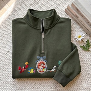 May include: An olive green quarter-zip sweatshirt featuring embroidered characters from The Little Mermaid, including Ariel in a pearl-framed cameo, Sebastian, Flounder, and Scuttle. The garment has a silver-toned metal zipper and ribbed cuffs.