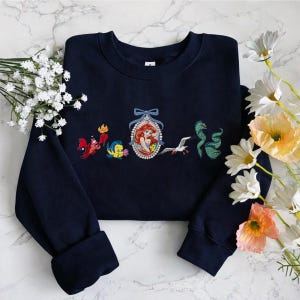 May include: A navy blue crewneck sweatshirt featuring an embroidered design of a mermaid in a pearl-framed oval, flanked by a red crab, a yellow and blue fish, a white seagull, and a green eel, all arranged horizontally across the chest.