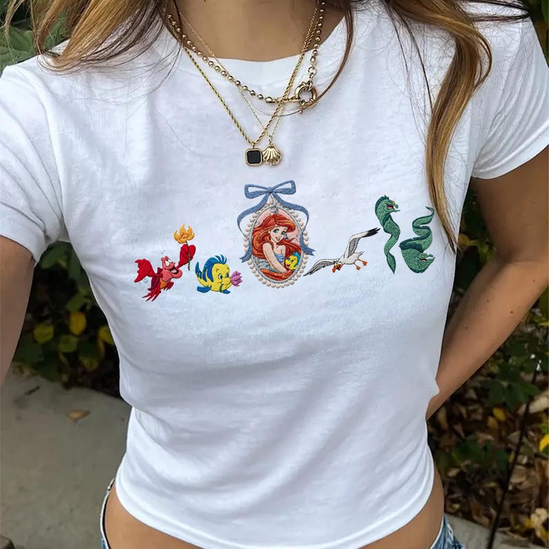 May include: A white short-sleeved t-shirt featuring a central embroidered graphic of a mermaid in an oval frame with a blue bow, flanked by a red crab, a yellow fish, a white seagull, and a green eel, all arranged in a horizontal line.