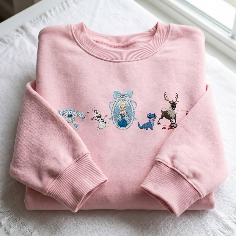 May include: A light pink crewneck sweatshirt featuring an embroidered design of five characters from the movie Frozen. The embroidery includes a white snow monster, a snowman, a blonde character in a blue frame, a blue lizard, and a brown reindeer.
