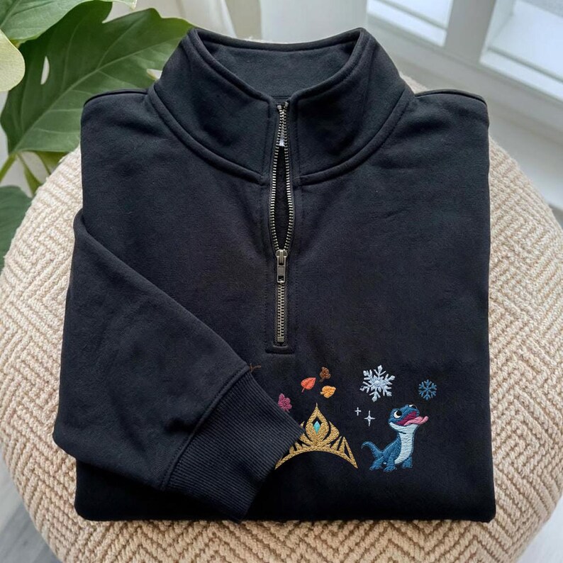 May include: A black quarter-zip sweatshirt featuring an embroidered design on the chest. The embroidery includes a gold crown, colorful autumn leaves, white snowflakes, and a small blue lizard character with its tongue sticking out.